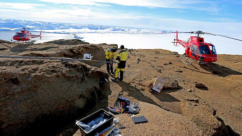 Expeditioners standing on a rocky surface with 2 helicopters in the background