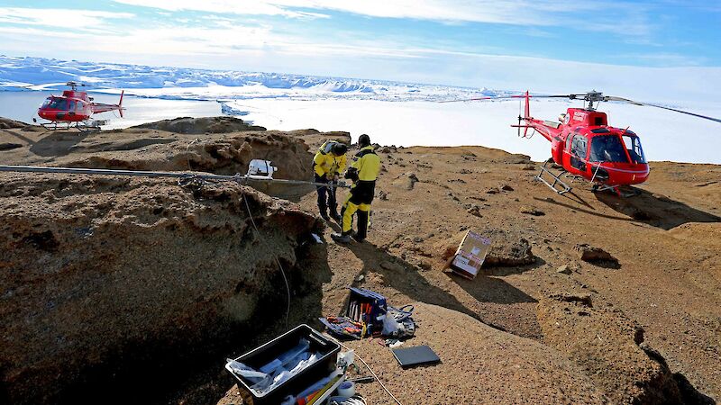 Expeditioners standing on a rocky surface with 2 helicopters in the background