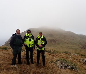 Three men stand on a barren hillside with mist in the background