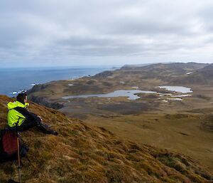 A person in yellow stands on a hillside and looks out at the view of lakes, ocean and wilderness