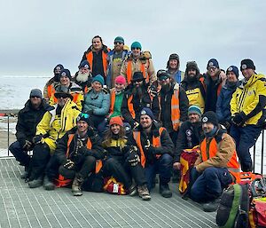 A group of smiling people stand on a steel deck and pose for a photograph - the sea-ice is in the background.