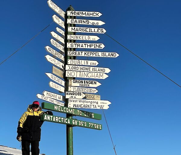 A smiling woman stands next to a Welcome to Davis Antarctica sign. The sign is adorned with street signs pointing to may locations on earth.