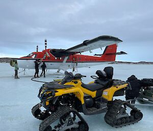 A yellow quad track bike in the foreground with a red and white aircraft in the background. People are refuelling the aircraft.