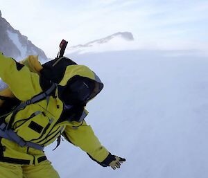 A person in yellow survival clothes mimics flying an aeroplane while standing in a windy snow-blown landscape.