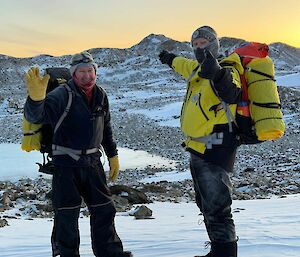 Two smiling men, waving at the camera, wearing backpacks hike through a snow-covered hilly landscape.