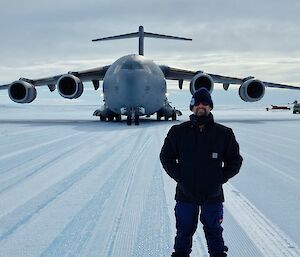 A man in black cold weather jackets is smiling at the camera. In the background is a large, four engined, cargo aircraft
