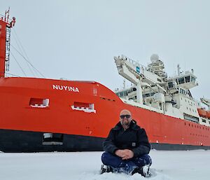 A man is sitting on the sea ice in front of a large red ice breaker