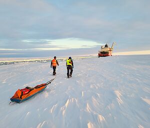 Two people are walking towards a large icebreaker in the distance. They are pulling a sledge with a generator and other equipment on it.