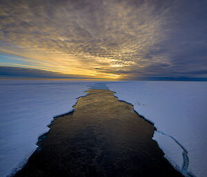 A broken path through the sea ice left by an icebreaker stretches into the distance. The sun is low on the horizon.