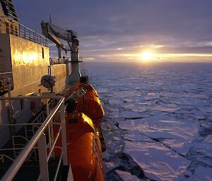 Looking from the side of an icebreaker, the sun is setting on the horizon over a sea covered by broken sea ice