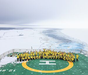 A large group of people in bright yellow cold weather jackets are posing for a group photo on the helicopter deck of an icebreaker ship