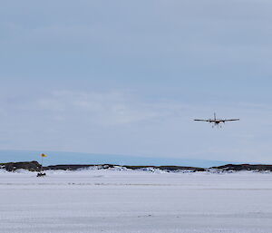 A twin engined light aircraft is coming towards the camera to land on the sea ice. There is a wind sock on the left of frame and a rocky island in the distance.