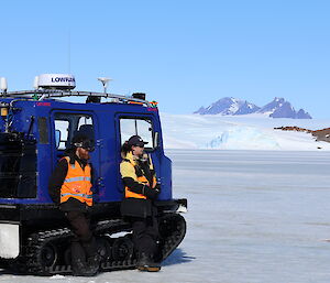 A woman and a man are leaning against a blue Hägglunds vehicle that is parked on the sea ice. In the distance is an ice plateau with a rocky mountain range on it