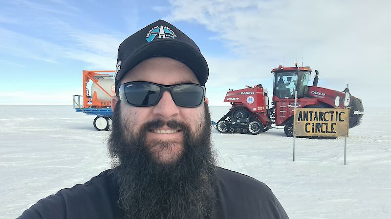 A man in black with a beard stands on ice in front of the Antarctic Circle sign, with a tractor in the background