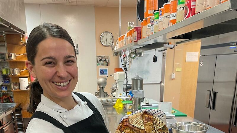 A smiling woman in a black apron stands in a kitchen holding a loaf of bread