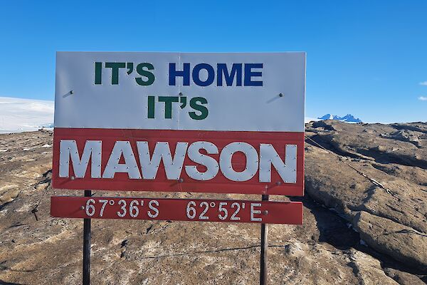 A sign saying "It's home, it's Mawson" against a rocky landscape with ice in the distance