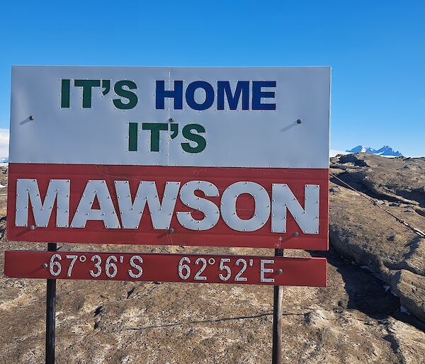 A sign saying "It's home, it's Mawson" against a rocky landscape with ice in the distance