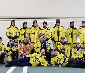 Thirteen men and two women, all wearing cold weather clothing, are posing for a group photo
