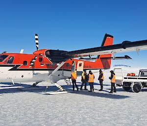 A red and white light aircraft is parked on the sea ice and a number of people are unloading items onto a utility