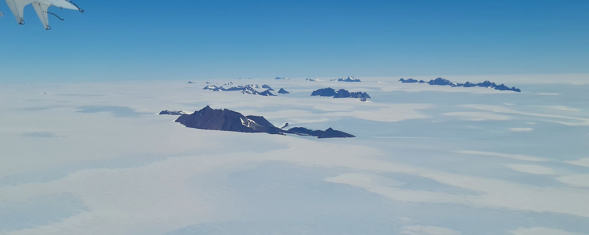 An aerial view of a number of rocky mountain ranges rising from the ice plateau