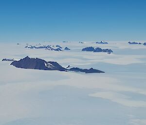 An aerial view of a number of rocky mountain ranges rising from the ice plateau