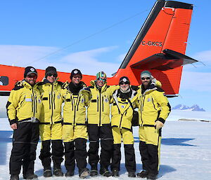 Six people wearing bright yellow cold weather jackets are posing for a photograph in front of a light aircraft that is parked on the ice