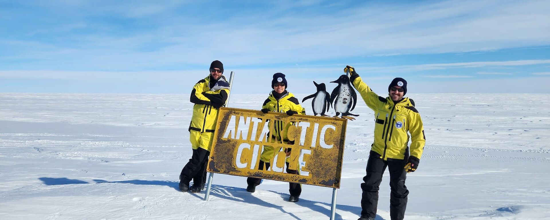 Three expeditioners standing on snow at the Antarctic Circle sign