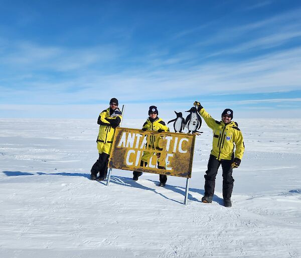 Three expeditioners standing on snow at the Antarctic Circle sign
