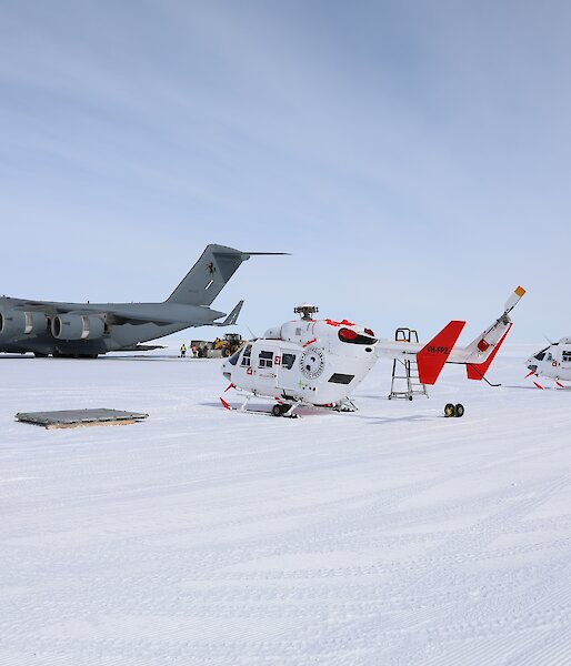 A C17 Globemaster sits on the ice with two helicopters in front of it.