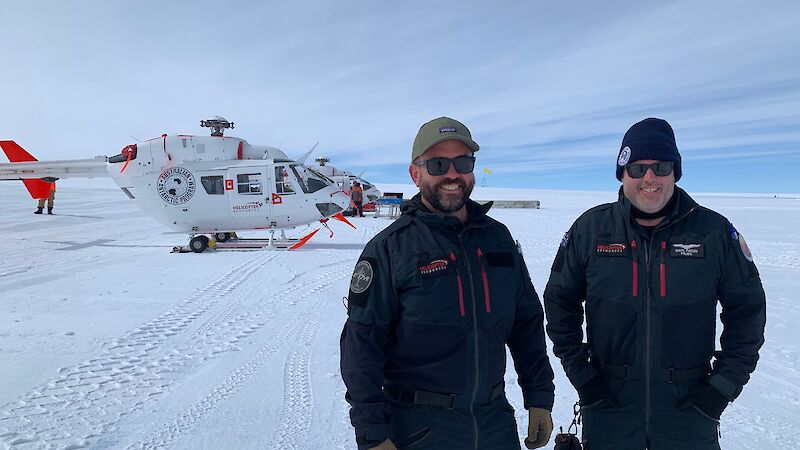 Two men in black alpine clothing in front of two helicopters, standing on snow