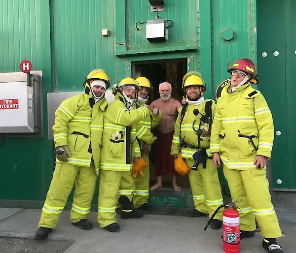 Five people in fire fighting uniform stand in front of a green building with a man smiling in the middle of them.
