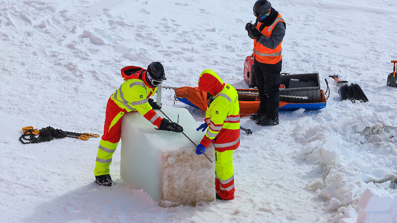 men in high vis use a massive saw to cut a block of ice