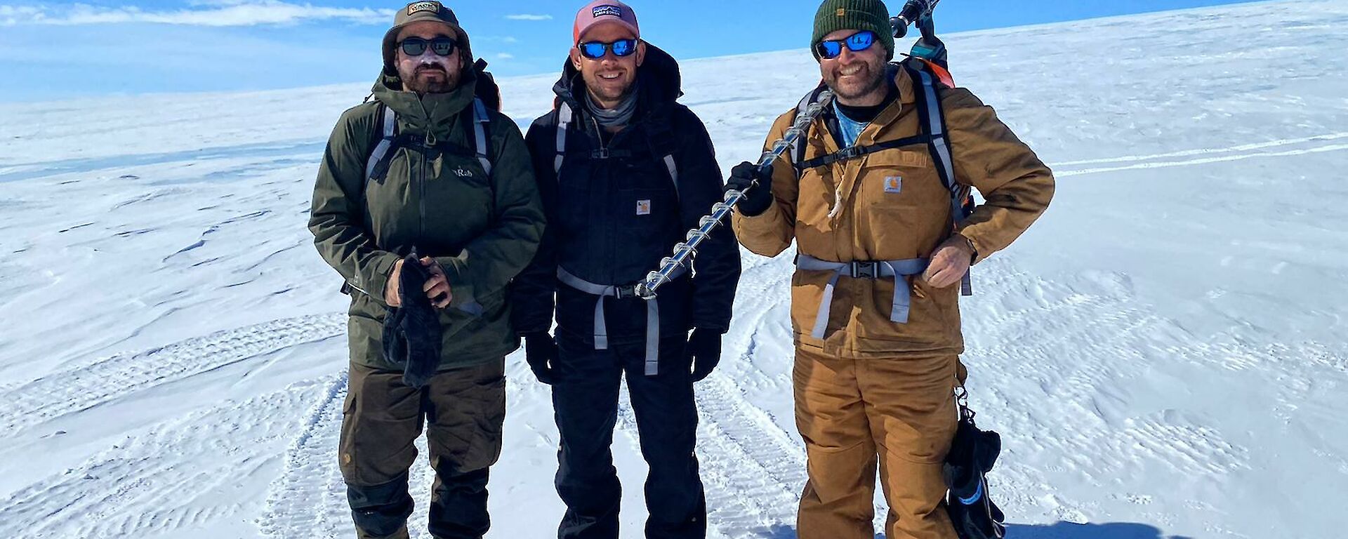 Three Antarctic expeditioners stand on snow holding an dice drill