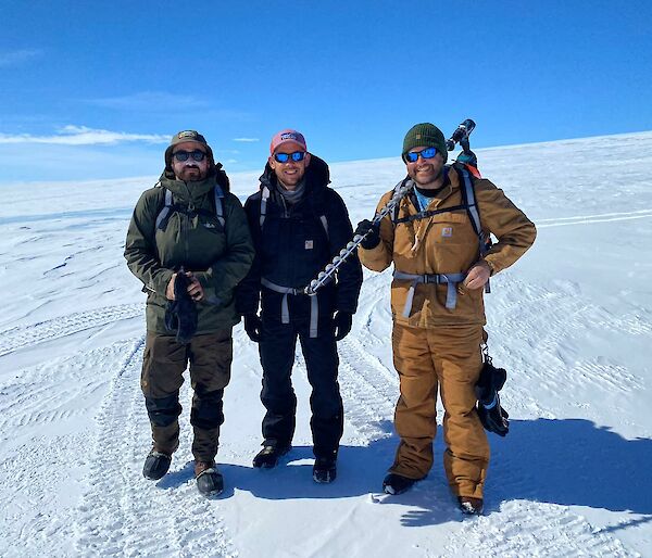 Three Antarctic expeditioners stand on snow holding an dice drill