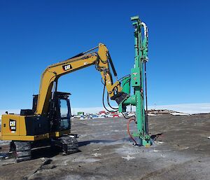 An excavator with drilling attachment is positioned on a rocky landscape. In the distance, multi coloured buildings of a station are visible.