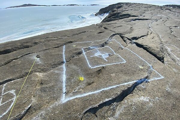 White paint markings on a rocky landscape depict a square hole to be cut. In the background is a frozen sea.