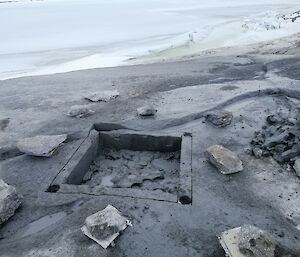 A square hole has been roughly cut out of the rock. The rocky landscape slopes away in the background to a frozen sea