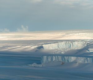 An ice plateau ends in cliffs above a frozen sea. Part of the plateau has carved off into an iceberg to the right of frame.