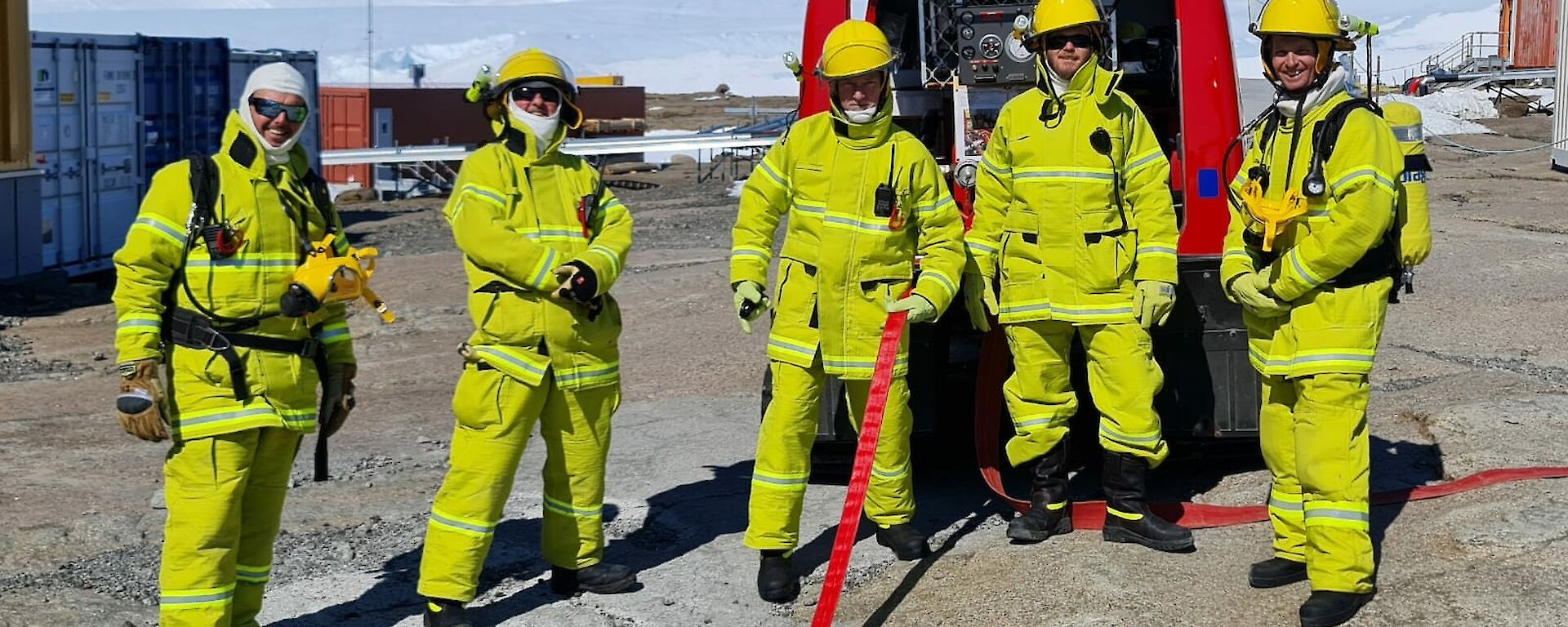 Five men are dressed in fire fighting clothing and standing in front of a red vehicle. A fire hose is extended from the vehicle.