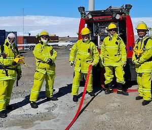 Five men are dressed in fire fighting clothing and standing in front of a red vehicle. A fire hose is extended from the vehicle.