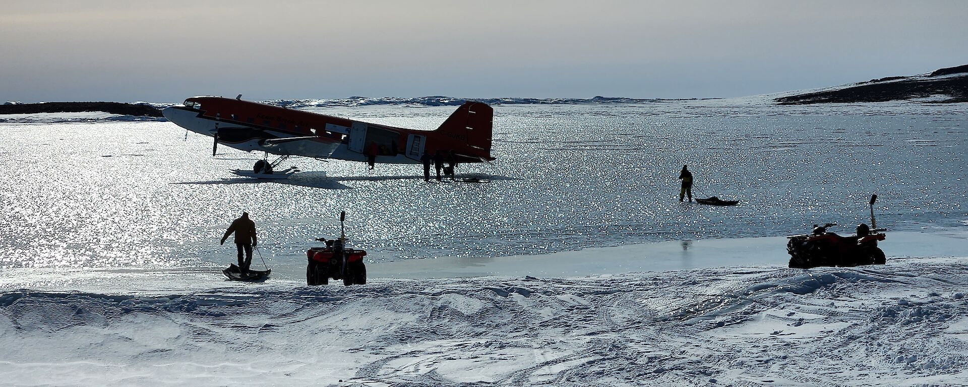 A red Basler plane sits on ice with people taking cargo on sleds away from from it.