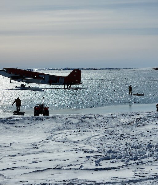 A red Basler plane sits on ice with people taking cargo on sleds away from from it.