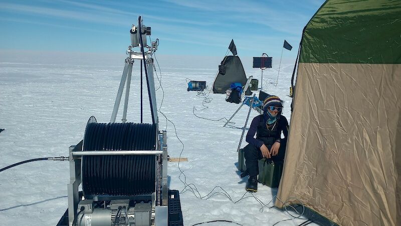 A person sits on the ice by a tent with a  big metal bog on their right.