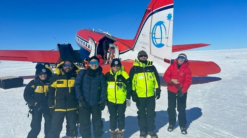 Six people stand on the ice in front of a red ski plane