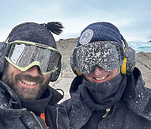 Two men are smiling at the camera. They are wearing goggles that are covered in splashed liquid making them hard to see through.