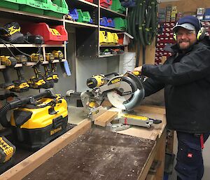 A man is smiling while working an a carpentry workshop. There are many battery powered drills and parts on the wall.