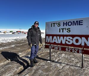 A man is standing in front of a sign that says "It's Home, It's Mawson". In the background are multi coloured station buildings against the ice.