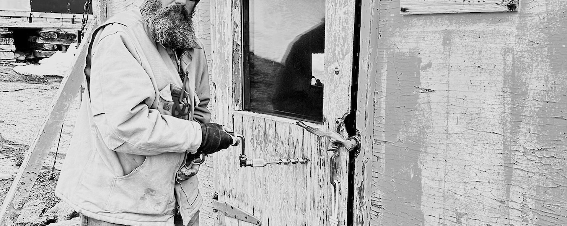 A black and white photograph of a man holding a hand drill against the door of an old, worn building.