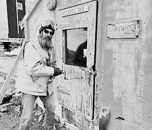 A black and white photograph of a man holding a hand drill against the door of an old, worn building.