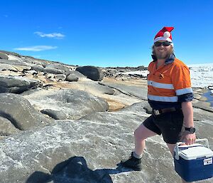 A man in a high visibility shirt and shorts is carrying an esky and walking across a rocky landscape. In the background are a number of Adélie penguins laying on the rocks